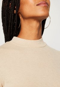 Haut beige côtelé avec un col montant, présentant une texture lisse et une coupe ajustée. De grandes boucles d'oreilles en or complètent ce look.