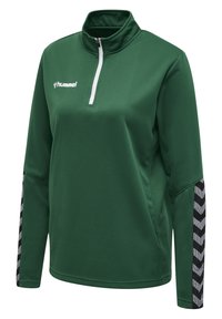Chaqueta de pullover verde con media cremallera, textura acanalada y patrón en chevron blanco y negro en las mangas. Logo de Hummel en el frontal.