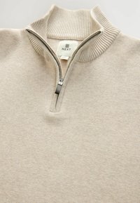 Pulover bej cu fermoar, având un guler înalt cu nervuri, o textură moale din tricot și un mic eticheta cu logo aproape de fermoar.