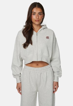 Kobieta o długich ciemnych włosach nosi jasnoszarą, skróconą bluzę z zamkiem i dopasowane spodnie dresowe na jednolitym tle.