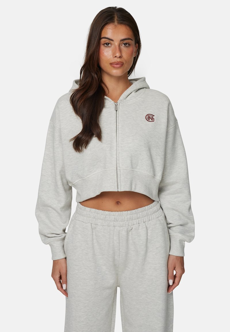 Kobieta o długich ciemnych włosach nosi jasnoszarą, skróconą bluzę z zamkiem i dopasowane spodnie dresowe na jednolitym tle.