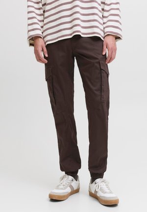 Personne portant un pantalon cargo marron foncé, des baskets blanches avec des accents beige et un haut à manches longues rayé beige et blanc.
