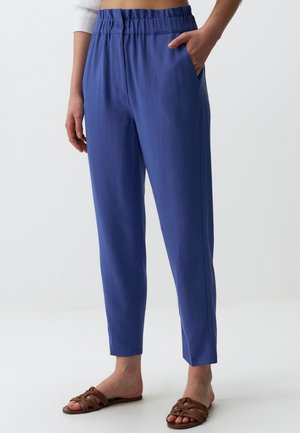 Femme portant un pantalon bleu taille haute, coupe décontractée avec une ceinture élastique et des sandales plates tressées marron, debout avec une main dans la poche.