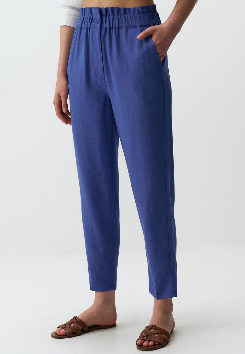Femme portant un pantalon bleu taille haute, coupe décontractée avec une ceinture élastique et des sandales plates tressées marron, debout avec une main dans la poche.