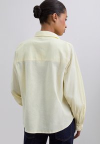Chemise longue à manches jaunes pâles avec une coupe décontractée, un col et des poignets élastiques. Présente une couture de yoke dans le dos et une texture douce.