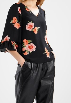 Blusa nera a fiori con motivi rosa e verdi; scollo a V, maniche corte con volants. Indossata con pantaloni in finta pelle nera, texture liscia.