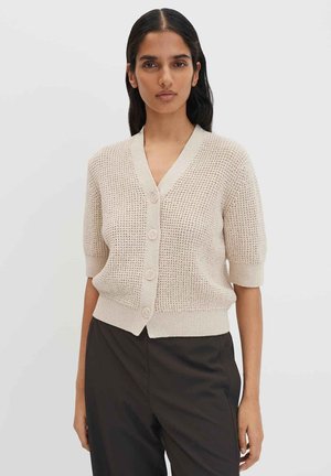 Donna che indossa un cardigan beige testurizzato a maniche corte con bottoni e pantaloni neri, in piedi su uno sfondo chiaro e uniforme.