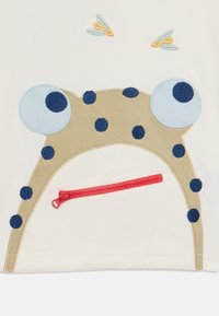 T-Shirt mit einem Froschdesign in Blau auf beigem Stoff und einer roten Reißverschlusstasche. Zwei gestickte Libellen über dem Design.