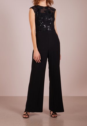 Kobieta w czarnym, bezrękawnym kombinezonie z cekinowym kwiatowym topem i szerokimi spodniami, w połączeniu z czarnymi sandałkami na paski.