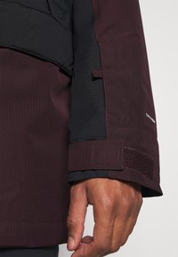 Primer plano de una mano que lleva una chaqueta impermeable de color burdeos oscuro y negro, con puño ajustable de Velcro y un pequeño detalle de lazo de tela.