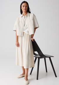 Chemisier court beige clair à manches courtes sur un haut blanc texturé, associé à une jupe fluide crème et des ballerines beiges. Chaise noire en arrière-plan.