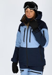 Ski jacket in navy and light blue, featuring a hood, zip pockets, and black accents. Paired with matching pants. Soft texture.