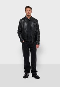 Chaqueta de cuero negra con cierre de cremallera, que presenta un cuello alzado y un ligero brillo. Combinada con pantalones oscuros y zapatos de cordones negros.