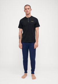 Polo preto de manga curta com um padrão texturizado, três botões no colarinho e logótipo amarelo "POLO". Combinado com umas calças de lazer azul-marinho.