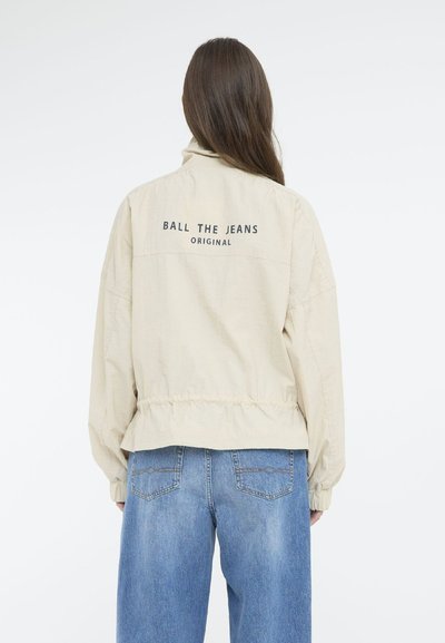 Veste beige avec un col montant, une taille élastique et une impression au dos "BALL THE JEANS ORIGINAL." Associée à un jean bleu à jambes larges.
