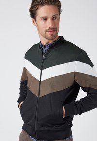 Giacca con zip caratterizzata da pannelli verde, bianco, marrone e nero in un design a zigzag. Realizzata in un tessuto liscio con tasche laterali e polsini a coste.