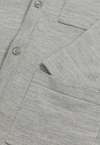 Hellgrauer Cardigan aus weichem Strickstoff, ausgestattet mit vier runden Knöpfen und einer aufgesetzten Tasche auf der Vorderseite. Subtile Textur sichtbar.