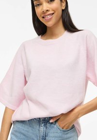 Oblečen svetlo roza ohlapen pulover iz mehkega pletenega blaga z okroglim izrezom in kratkimi, širokimi rokavi, kombiniran z visokim pasom denim hlač.