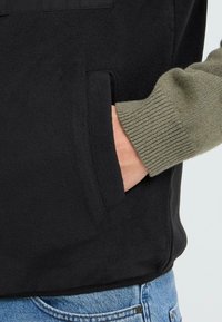 Schwarze Fleecejacke mit olivgrünen gerippten Bündchen und Taschendetails, die eine weiche Textur und ein lässiges Design präsentieren.