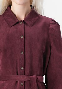 Bordeauxfarbener Kordkleid mit Kragen, durchgehender Knopfleiste und taillierter, gerippter Textur sowie dezenten Stickdetails.