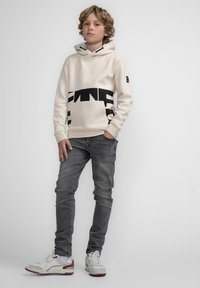 Felpa crema con un design grafico nero, jeans grigi e sneaker bianchi con dettagli rossi. La felpa presenta una tasca frontale e un cordino.