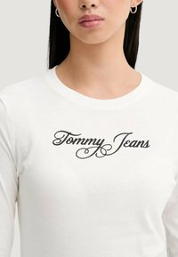 Biały top z długim rękawem i okrągłym dekoltem. Na piersi czarny, kursywny napis "Tommy Jeans". Miękka, gładka faktura materiału.