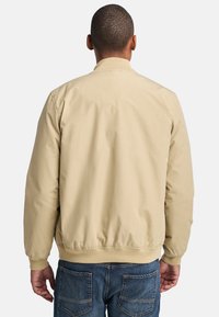 Chaqueta bomber beige con una textura suave, puños y cinturilla elásticos, y un cuello simple, combinada con unos jeans de mezclilla azules.