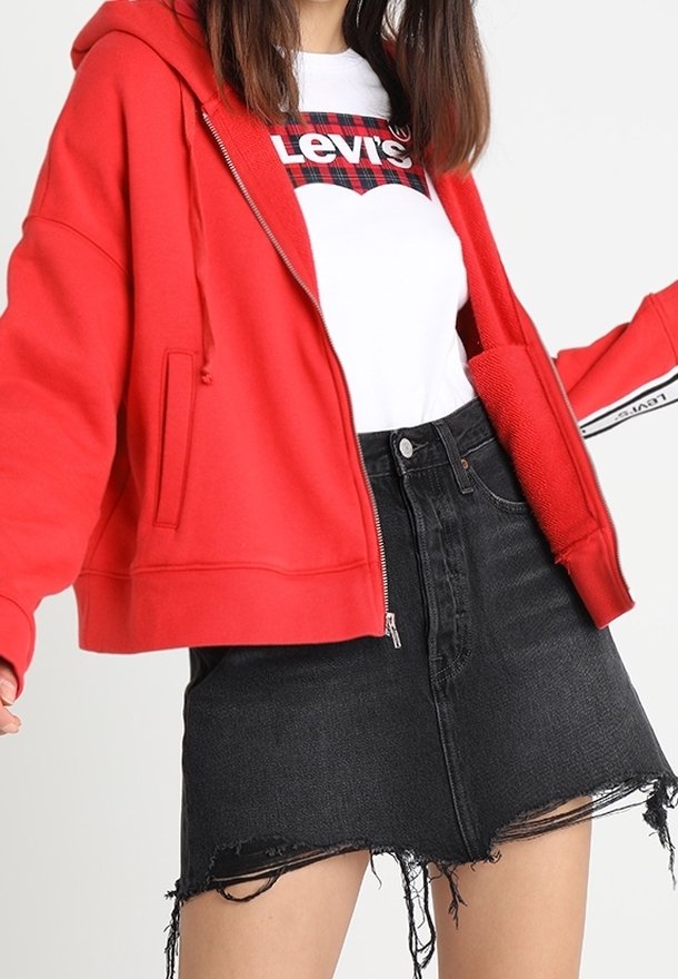 Femme portant un sweat à capuche rouge zippé, un t-shirt blanc Levi's et une mini-jupe en jean noire déchirée, posant avec les bras légèrement écartés.