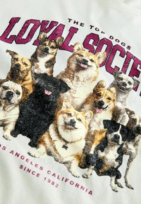 T-shirt graphique blanc présentant un imprime de différents chiens, y compris des Corgis et un chien noir, avec des accents scintillants et le texte "Société Loyale."