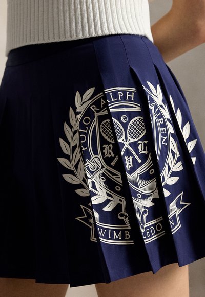 Ναυτικό πλισέ φόρεμα με λευκό σχέδιο έμβλημα Ralph Lauren Wimbledon με θέμα το τένις, φορεμένο με λευκό ριμπ μπλουζάκι.