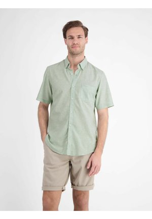 Man draagt een lichtgroen overhemd met korte mouwen en beige opgerolde korte broek, staand tegen een effen witte achtergrond.