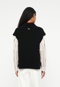 Sweater sem mangas preto com textura canelada, gola alta e fendas laterais, apresentando um pequeno detalhe de logotipo nas costas. Estilizado sobre uma camisa listrada.