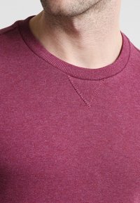 Närbild av en persons nacke och övre bröst som bär en vinröd rundhalsad tröja med ribbad krage och sömdetaljer.