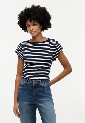 Vrouw met krullend haar staat, gekleed in een marineblauw-wit gestreept T-shirt met korte mouwen, gouden knopen en een high-waisted blauwe spijkerbroek, op een witte achtergrond.