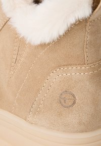 Beiger Wildleder-Stiefelette mit weißem Fellfutter. Verfügt über Steppnähte und ein rundes Logo an der Seite. Strukturierte Oberfläche und weiche Akzente.