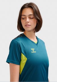 Tealfärgad sporttröja med gröna detaljer, korta ärmar och V-ringning. Har texturerad detalj på axeln och en logotyp framtill.