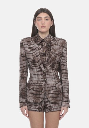 Donna con lunghi capelli scuri che indossa un completo coordinato con blazer, camicia e pantaloncini marroni e beige a motivo, con bottoni dorati, in piedi davanti a uno sfondo bianco.