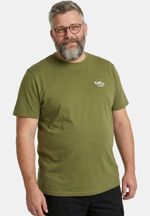 Brodaty mężczyzna w okularach, ubrany w oliwkowo zielony T-shirt z małym białym logo ryby na piersi, stojący na tle jednolitego jasnego tła.