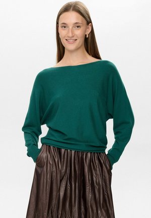 Pullover verde in maglia morbida e ampia, con scollo largo e maniche lunghe, abbinato a una gonna plissettata marrone. Texture soffice, design casual.