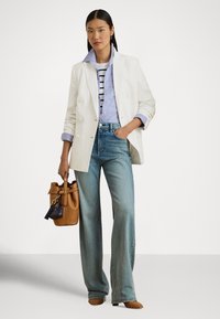 Γυναίκα που φοράει λευκό σακάκι πάνω από ριγέ πουλόβερ και μπλε πουκάμισο, τζιν με φαρδιά πόδια, καφέ τσάντα και καφέ παπούτσια με τακούνι, στέκεται με το ένα χέρι στην τσέπη.