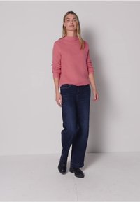 Pull côtelé rose à col haut, associé à un jean large bleu foncé et des chaussures noires. Un fond uni met en valeur la tenue.