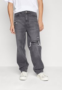 Šedé džínové kalhoty s volnějším střihem, s trhlinami nad koleny a jemným vyblednutím. Nošeny s bílou košilí a bílými teniskami.