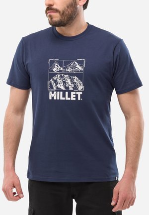 Homme portant un t-shirt bleu marine avec des illustrations de montagnes blanches et le texte "MILLET" sur le devant, debout devant un fond blanc.