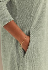 Grøn ribstrikket kjole med sidelomme, kombineret med et armbånd af guldkæde. Fokus på blød tekstur og afslappet design.