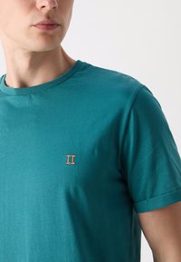 Teal bombažna majica s crew izrezom, zavitimi rokavi in majhnim oranžnim vezenim "II" na prsih. Gladka tekstura, sproščen kroj.
