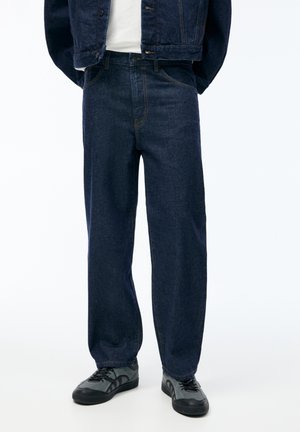 Personne portant un jean droit bleu marine associé à des baskets noires et grises ainsi qu'une veste en denim foncé assortie.