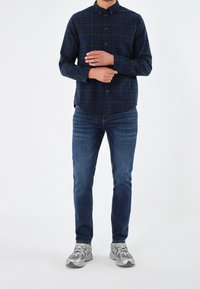 Marinblå rutig skjorta med gröna accenter, kombinerad med blå jeans och grå skor; har långa ärmar och rundad fåll.