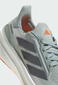 Zapatilla Ultraboost de Adidas en gris claro con rayas gris oscuro, logo naranja en la lengüeta y suela blanca texturizada con borde naranja en la parte inferior.