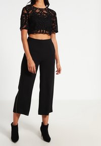 Sort cropped top med gennemsigtig blondetop og blomstermønster, parret med højhårede sorte culottes, der har lynlåsdetaile på siden.