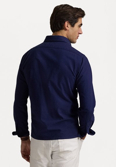 Camisa de manga larga azul marino con cuello abotonado, que presenta una textura suave y puños enrollados, combinada con pantalones cortos de color claro.